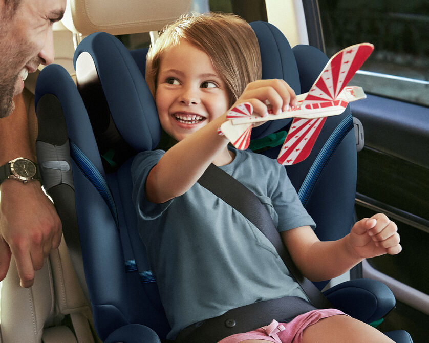 A child sitting in a car seat holding a toy airplane, smiling.