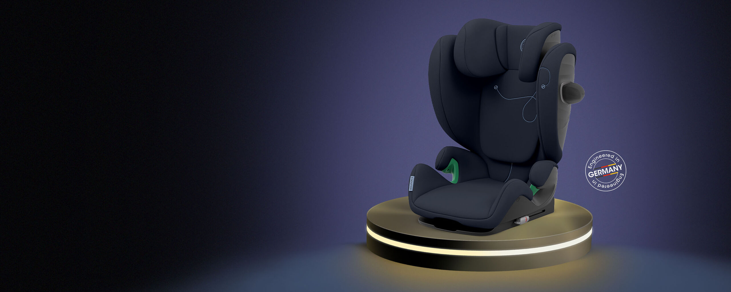 A modern black child car seat on an illuminated platform, with a "Engineered in Germany" seal next to it.