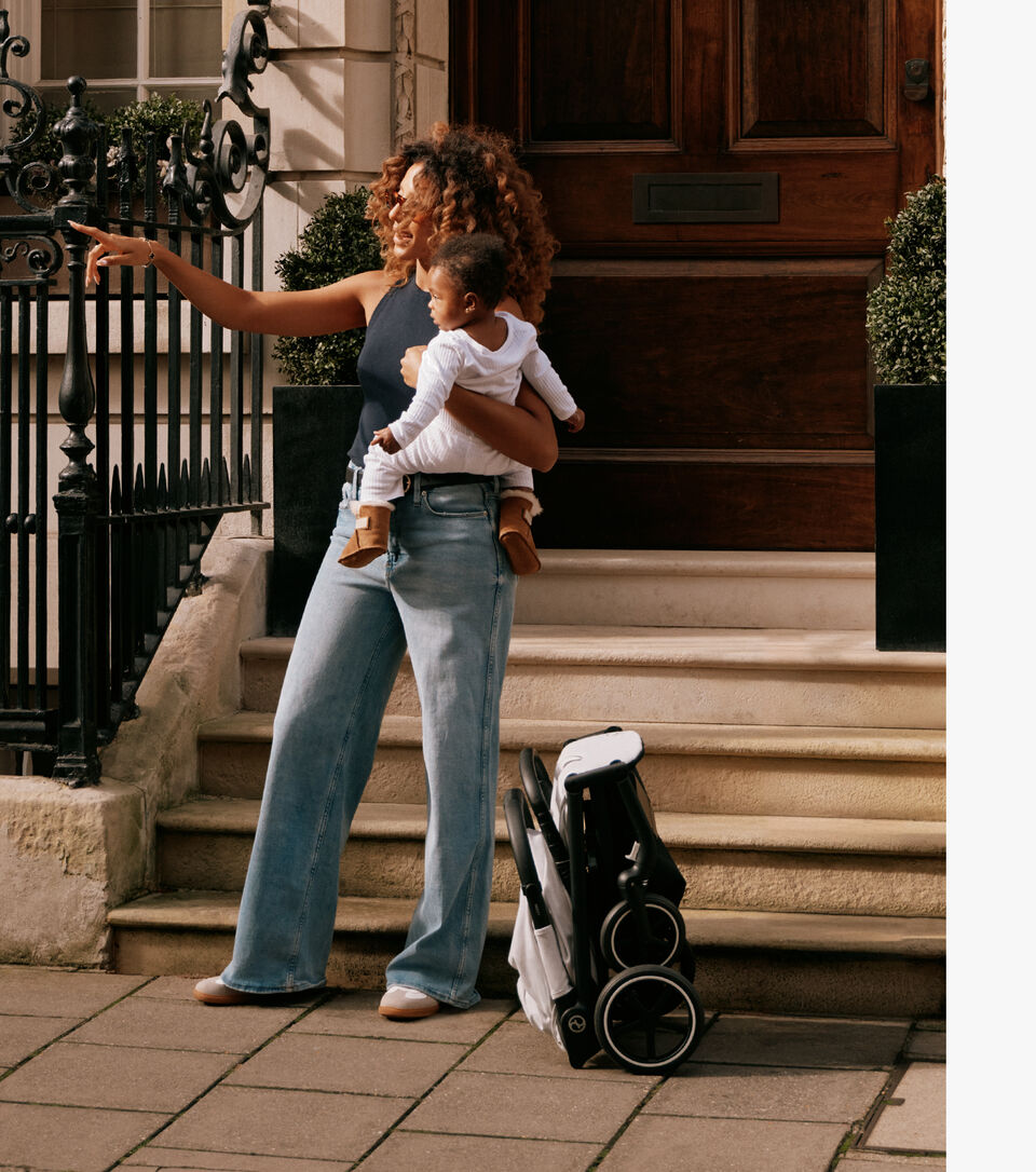 A woman with curly hair holds a baby in her arms while standing on steps next to a folded Beezy lightweight compact stroller.