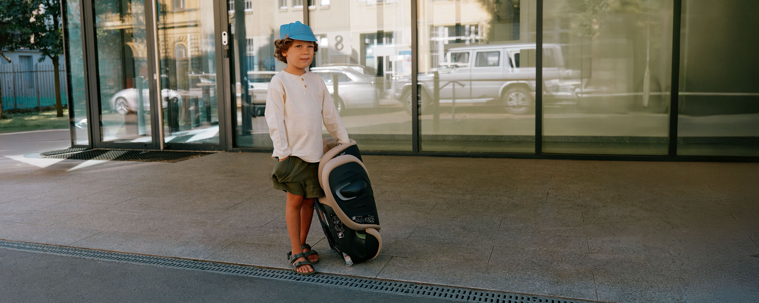 A child wearing a blue cap stands outside holding a car seat.