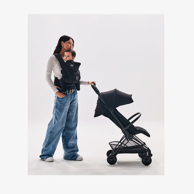 A woman carries a baby in a black baby carrier and stands next to a folded pushchair.