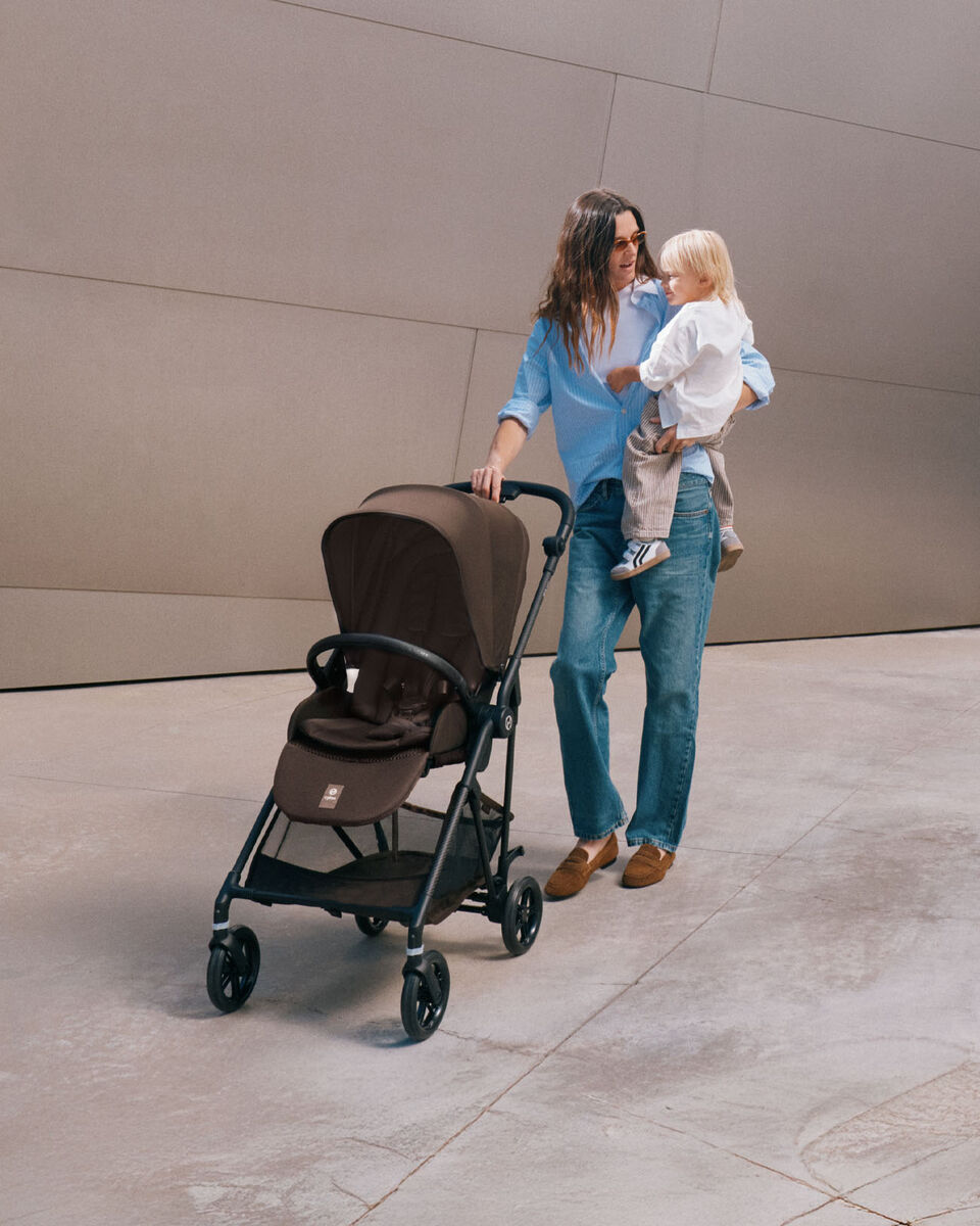 Una mamma spinge il passeggino Melio Carbon tenendo in braccio il suo bambino.