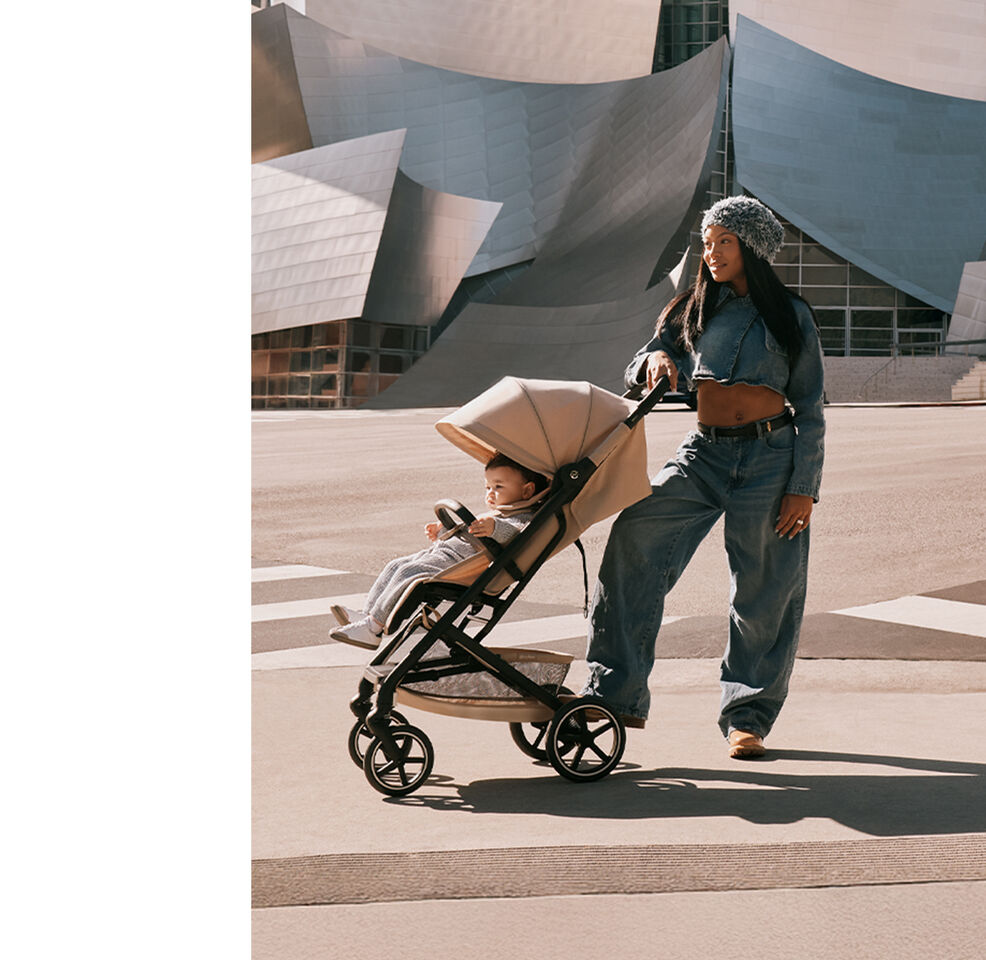 Une femme en denim et portant un chapeau, une main et un pied pos&eacute;s sur sa poussette compacte Beezy. Son b&eacute;b&eacute; est assis &agrave; l&rsquo;int&eacute;rieur de la poussette compacte.