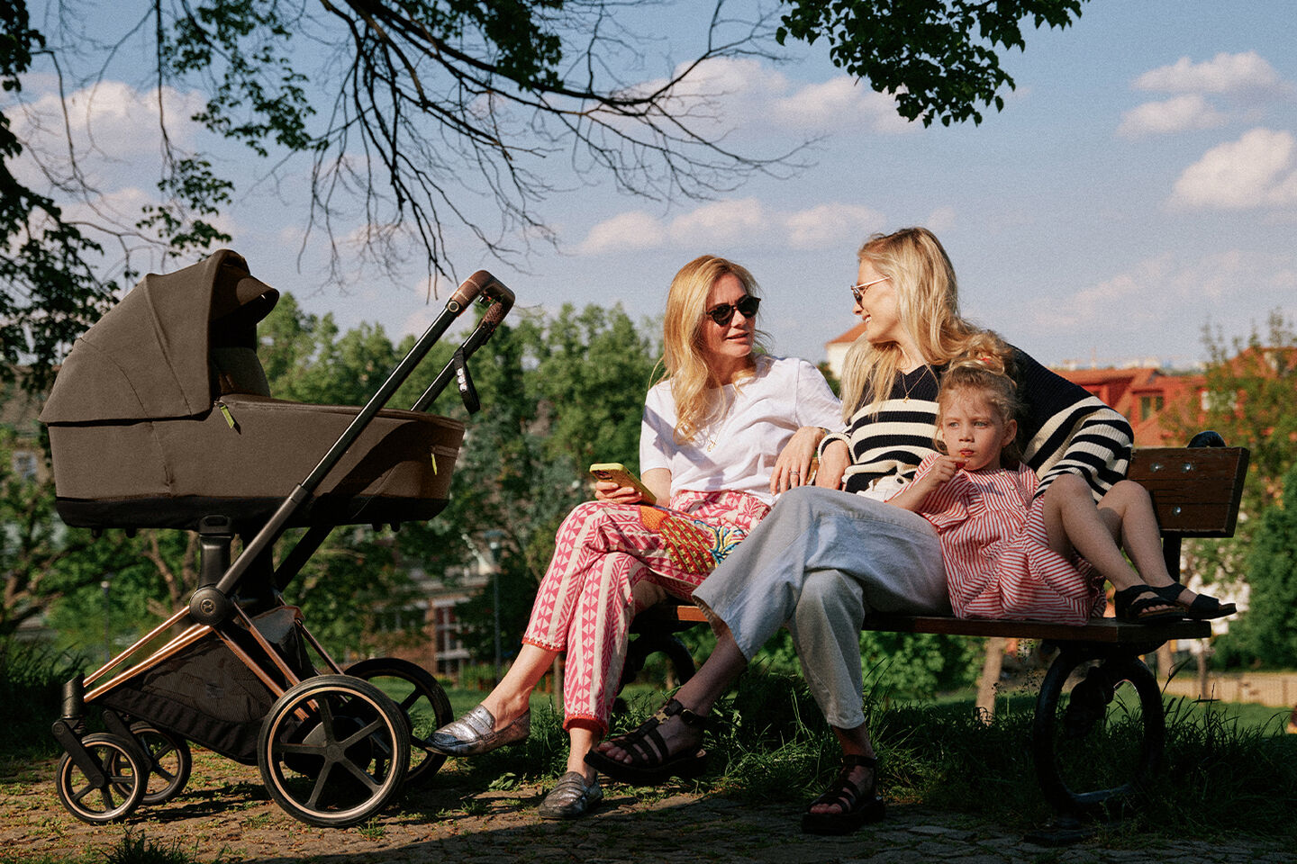 Two women and a small child sitting on a park bench next to a pram, surrounded by trees and blue sky.