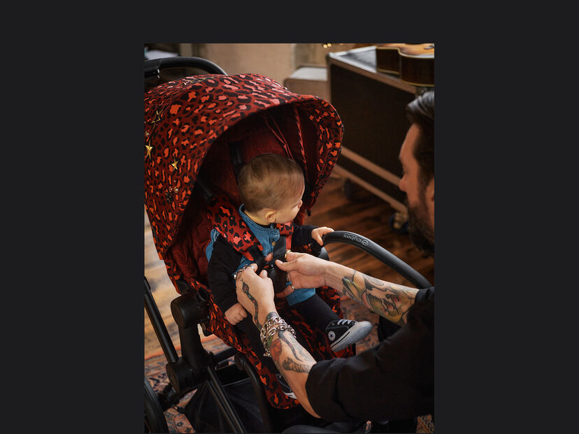 Ein Mann schnallt ein Baby in einem CYBEX Rockstar Kinderwagen mit rotem Leopardenmuster an.