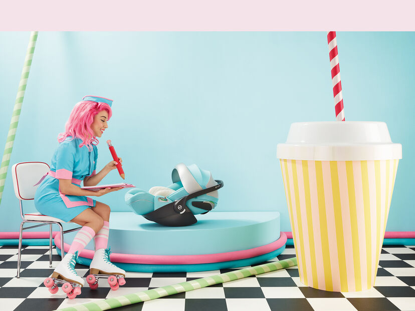 Žena v retro oblečen&iacute; s růžov&yacute;mi vlasy a kolečkov&yacute;mi bruslemi sed&iacute; na židli a drž&iacute; velk&eacute; červen&eacute; pero. Vedle n&iacute; je na podstavci autosedačka by Jeremy Scott Wings. V popřed&iacute; je vidět nadměrně velk&yacute; pruhovan&yacute; kel&iacute;mek s brčkem.