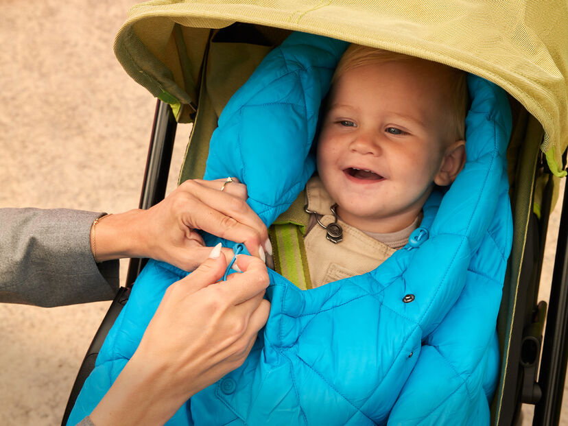 A smiling toddler sits in a pushchair, wrapped in a blue blanket, as an adult hand adjusts the blanket.