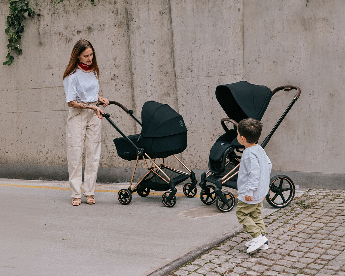 A woman pushes a pram with a toddler inside along a pavement.