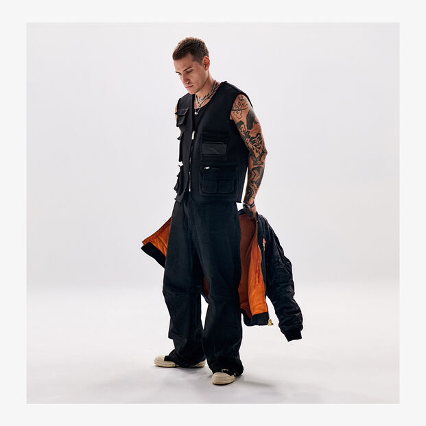 A man in black clothing and a vest with pockets, holding a jacket with orange lining.