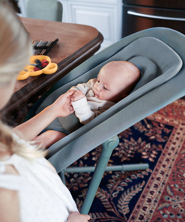 A baby lies in a grey baby seat, holding a woman's hand in a cosy room with a carpet and a table with a fruit bowl.