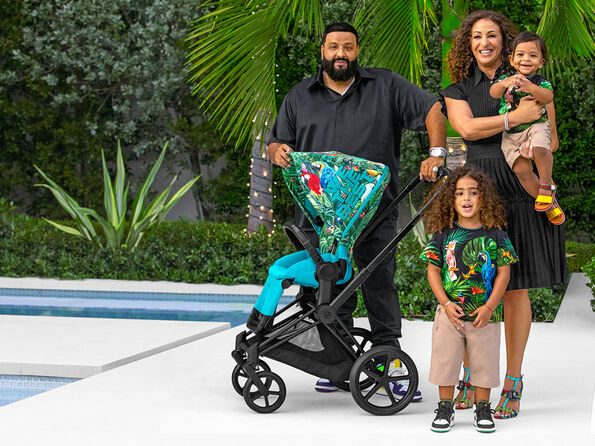 DJ Khaled with his family in a tropical look next to a Cybex pushchair with parrot design, taken in a garden by the pool