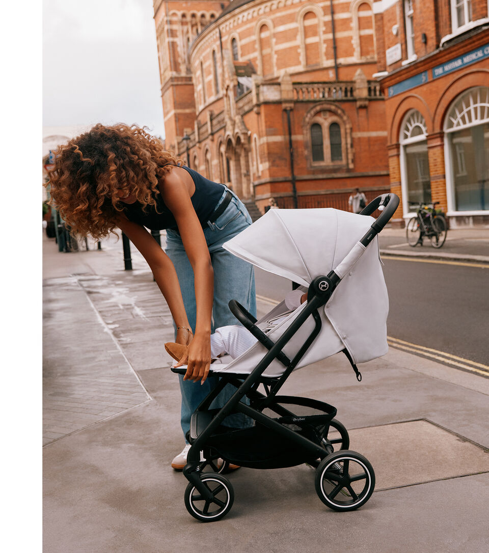 A woman with curly hair holds a baby in a white outfit while standing next to a Beezy lightweight compact stroller on a street.