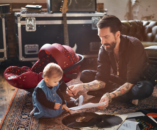 A tattooed man sits on a carpet showing a piece of paper to a toddler. Next to them is a red leopard-print child seat.