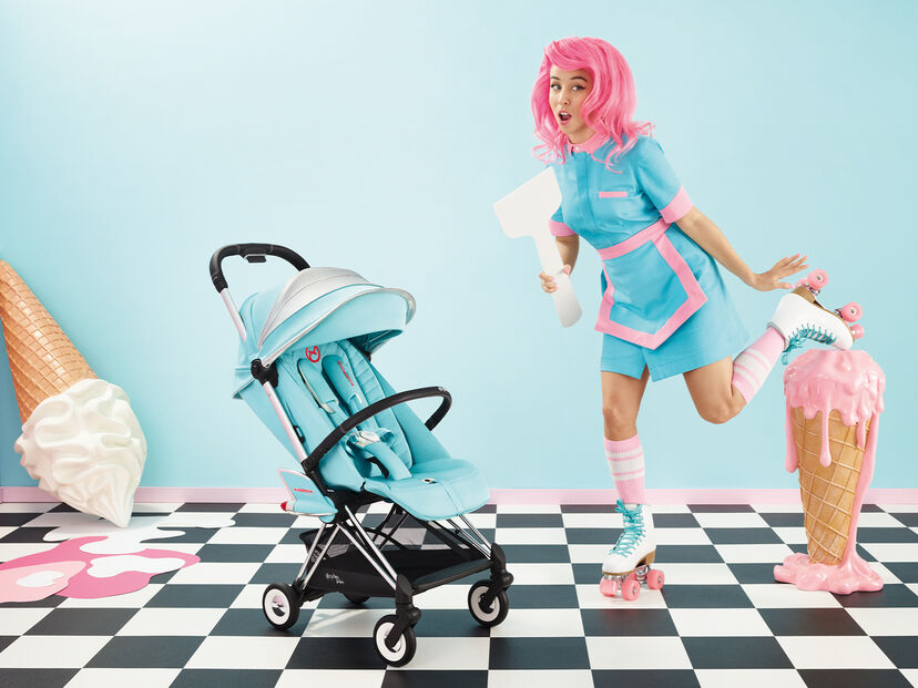 A turquoise stroller on a black and white chequered floor, next to a woman with pink hair in a turquoise dress on roller skates, surrounded by oversized ice cream cones.
