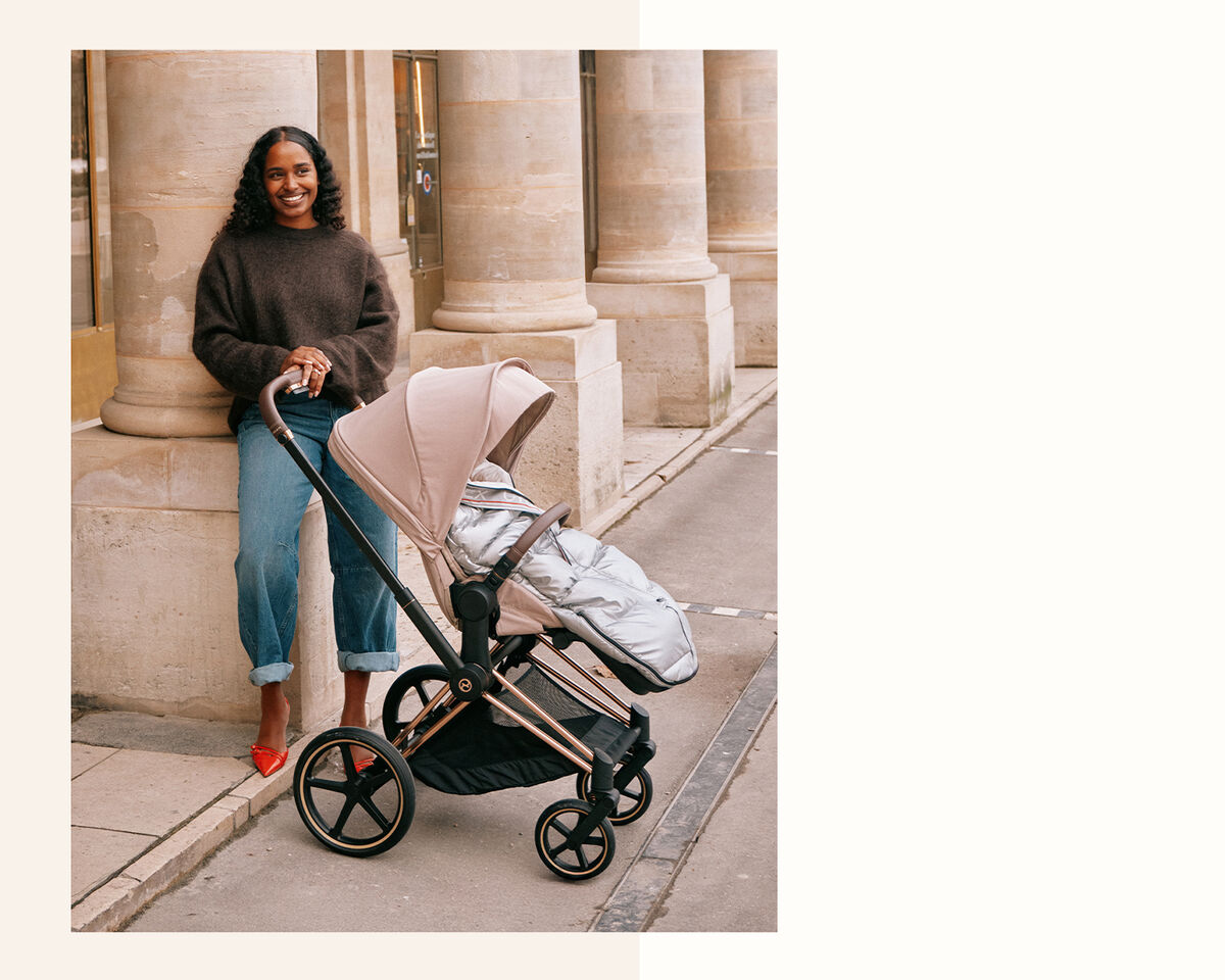 A woman with curly hair stands by a column, smiling while next to a modern stroller featuring a silver footmuff.