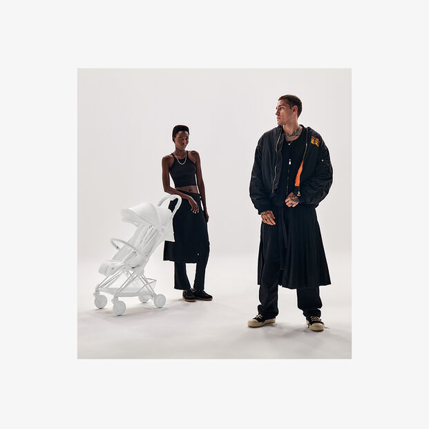 Two people in stylish black clothing stand next to a white pram in a minimalist studio.