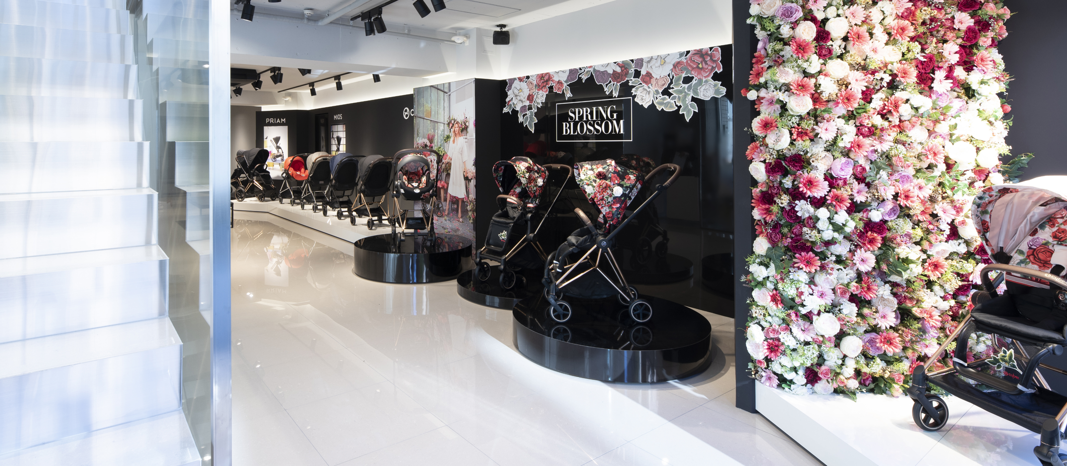 A stylish showroom featuring prams with floral designs, surrounded by a wall of colourful flowers.