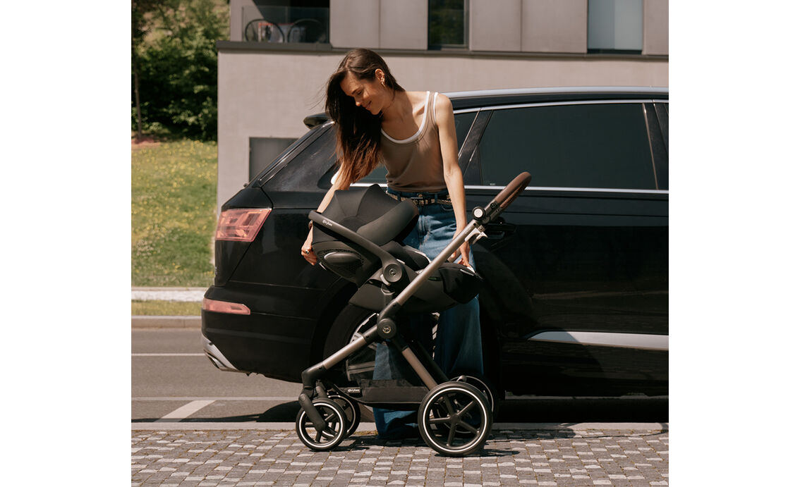 En brunh&aring;rig kvinna st&aring;r bredvid en svart SUV och lutar sig &ouml;ver ramen p&aring; en barnvagnsram fr&aring;n CYBEX med ett babyskydd f&auml;st. Suffletten p&aring; s&auml;tet &auml;r nedf&auml;lld.