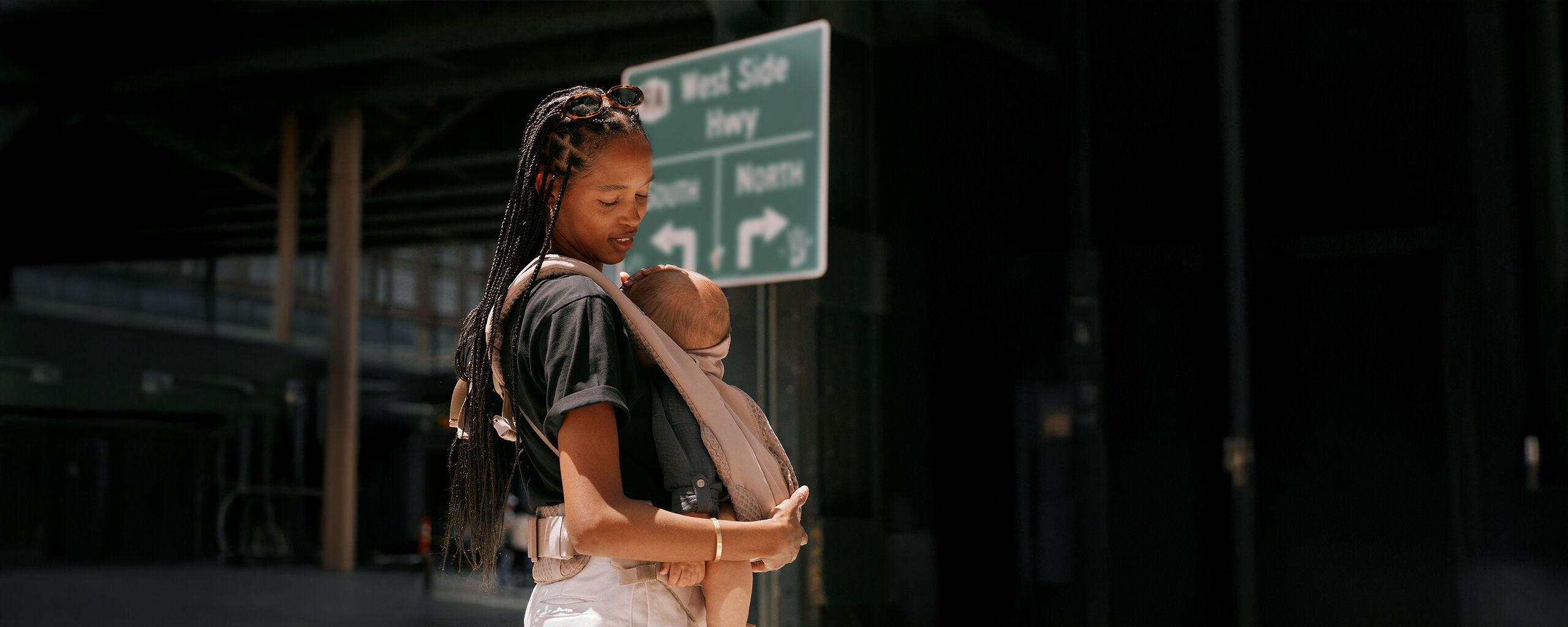 Een vrouw met vlechten draagt een baby in een Coya-draagzak naast een verkeersbord.