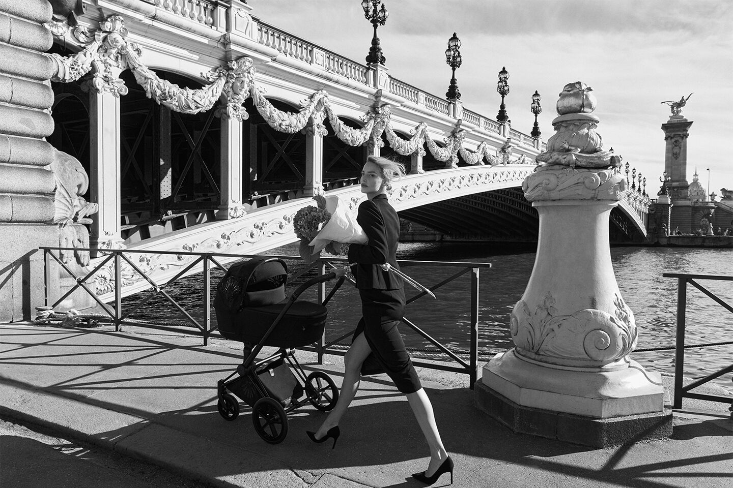 A stylish woman in an elegant black outfit pushes a pram across a bridge in Paris, holding a bouquet of flowers.