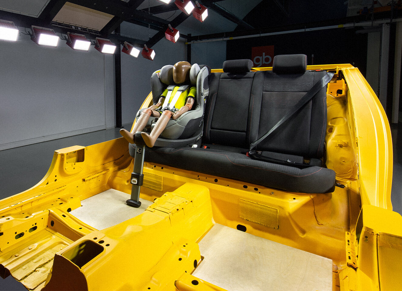 A crash test dummy is seated in a child car seat on the backseat of a yellow car frame.