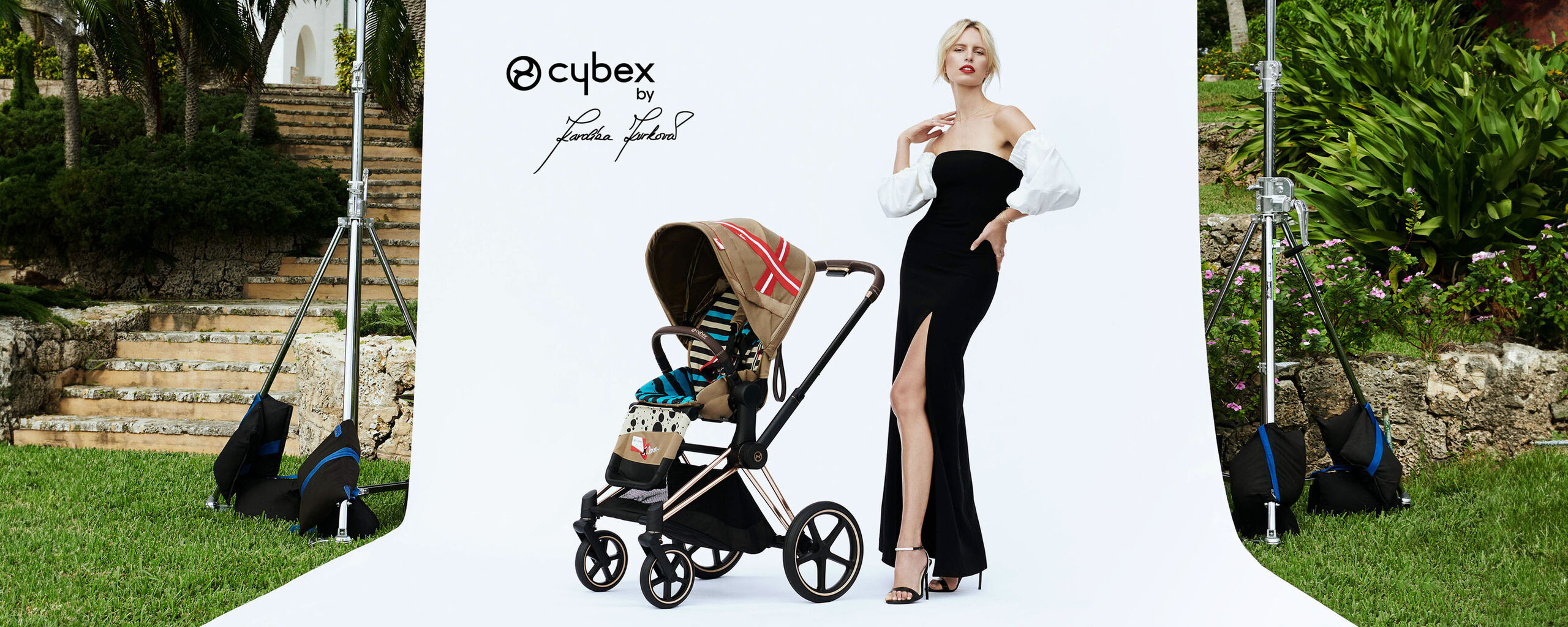 Elegantn&iacute; koč&aacute;rek Cybex by Karolina Kurkova vedle osoby v čern&yacute;ch &scaron;atech s b&iacute;l&yacute;mi ruk&aacute;vy na b&iacute;l&eacute;m pozad&iacute;.