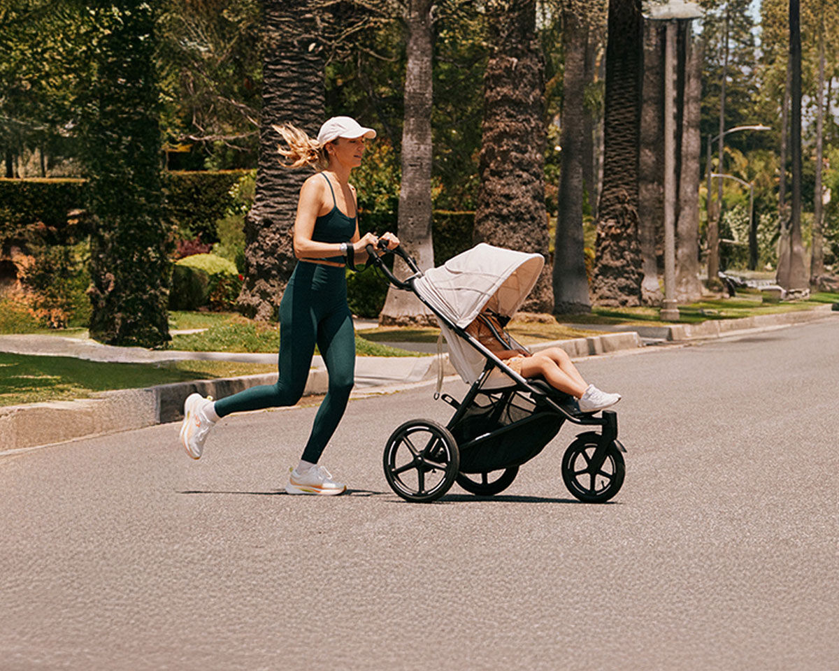 Woman jogging on a promenade with a child trailer, cityscape in the background.