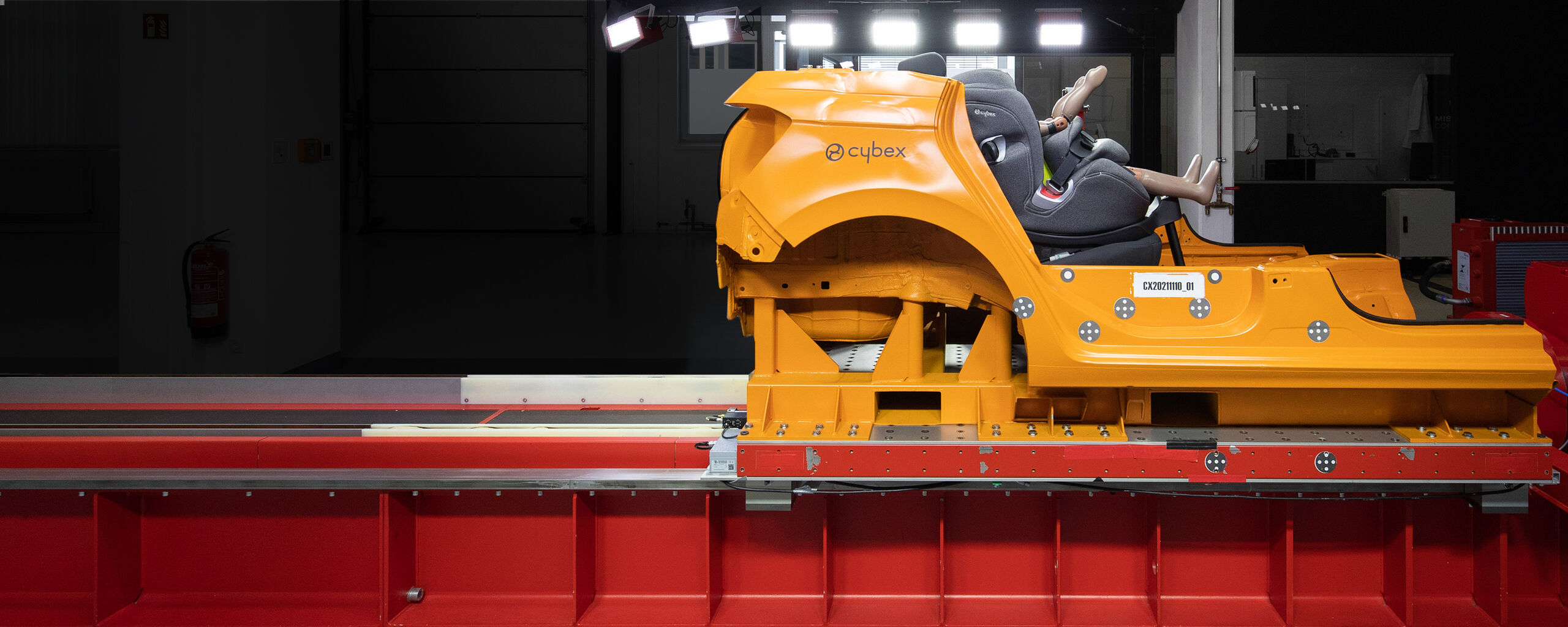 Ein orangefarbener Crashtest-Schlitten mit einem Cybex-Kindersitz und einer Dummy-Puppe in einer Testeinrichtung.