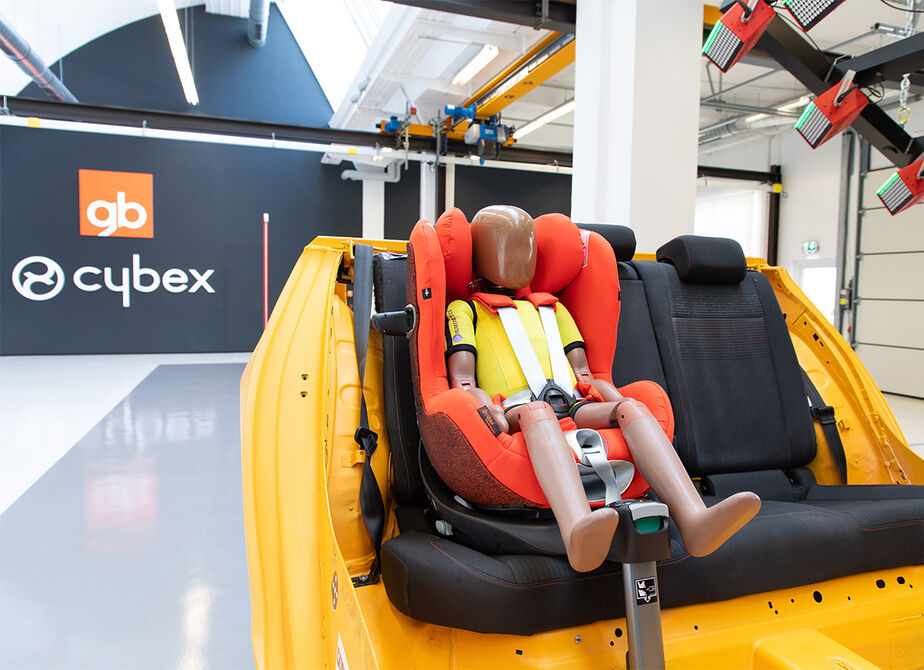 A crash test dummy seated in an orange child car seat within a vehicle interior.