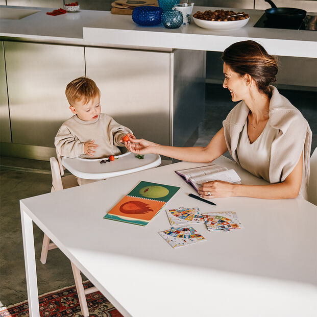 A toddler sits in a Click and Fold high chair playing with toys as a woman hands him something.