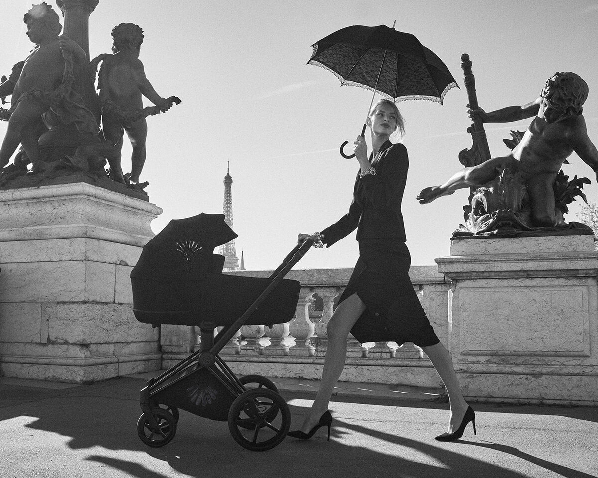 An elegant woman in a black dress and high heels pushes a La Parisienne pram on a Parisian bridge while holding a parasol. The Eiffel Tower is visible in the background.