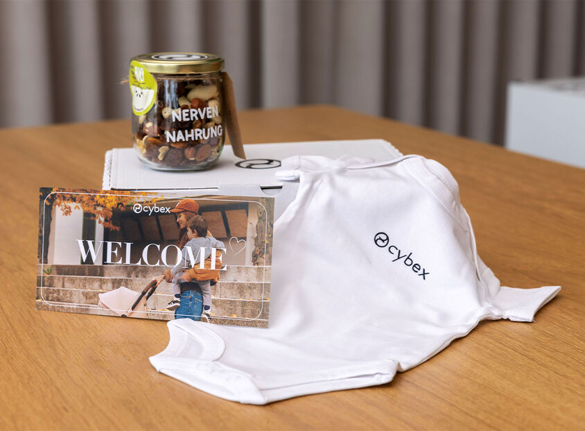 A baby onesie, a jar of nut trail mix, and a card reading &ldquo;Welcome&rdquo; arranged on a wooden table.