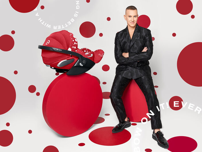 A man in a black suit sits on a red pedestal next to a red Cybex car seat with white polka dots.