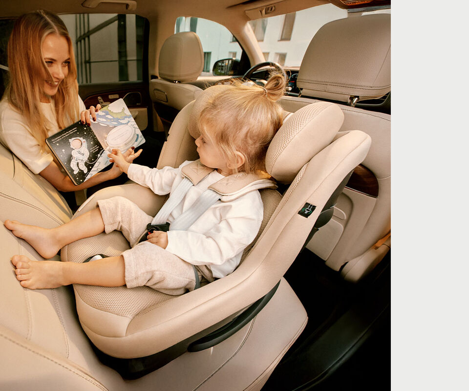 Een vrouw zit met haar dochter achter in de auto. De dochter zit in een Sirona G3 autostoel die naar achteren is gericht, en kijkt naar een prentenboek dat haar moeder haar laat zien.