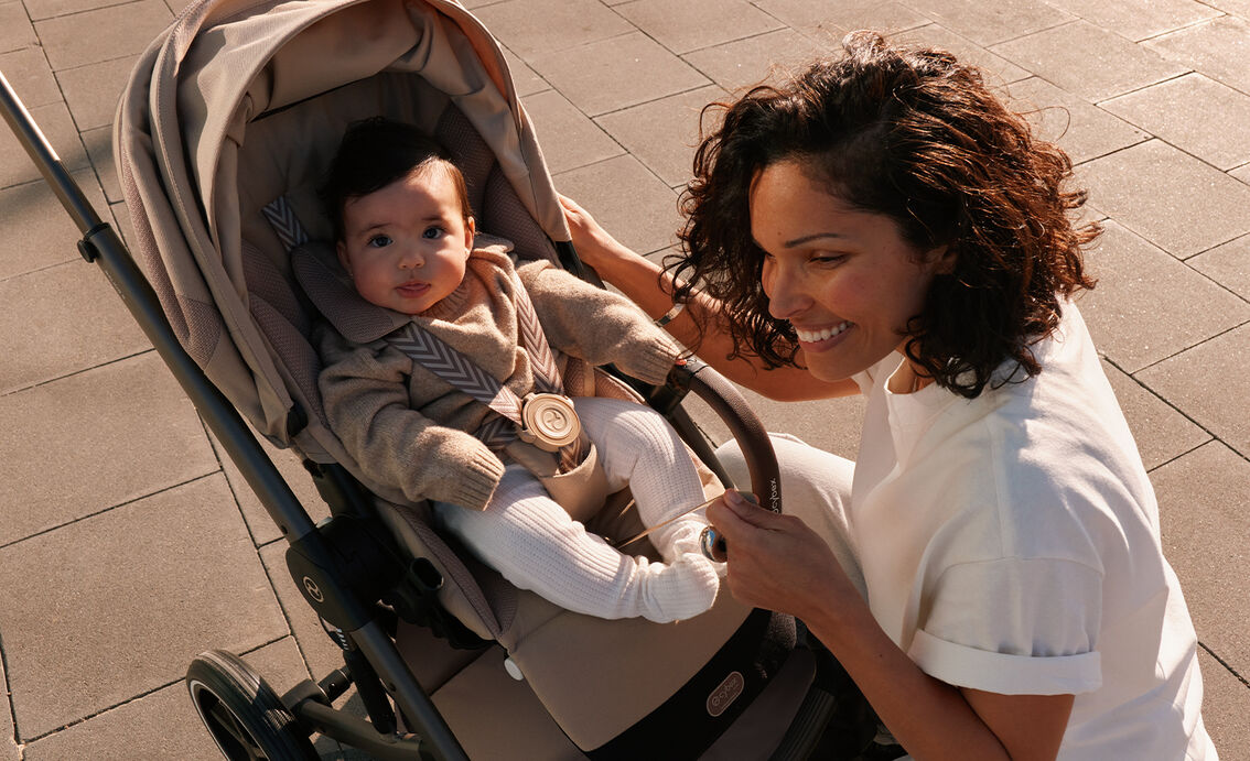 Eine Mutter kniet vor ihrem Balios Kinderwagen. Ihr Kind sitzt darin und schaut direkt in die Kamera.