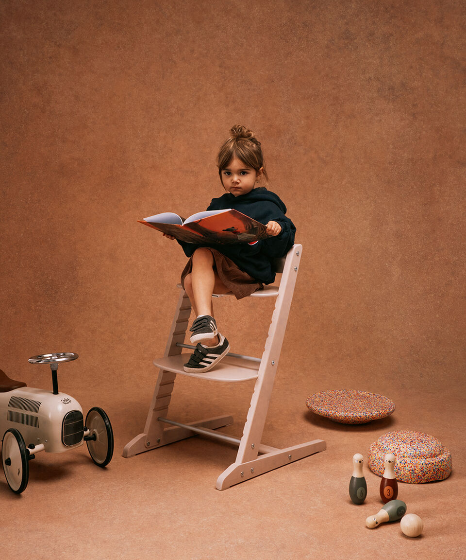 Little girl sitting crossed legged on a CYBEX Iris chair, holding a book in her hand, toys all around her.