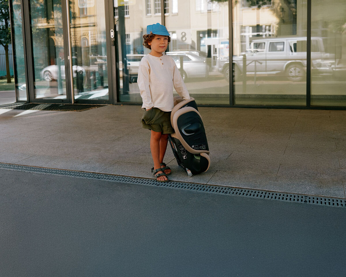 A small boy standing in front of a building with all glass windows. Next to him, a folded up Solution G2 car seat.
