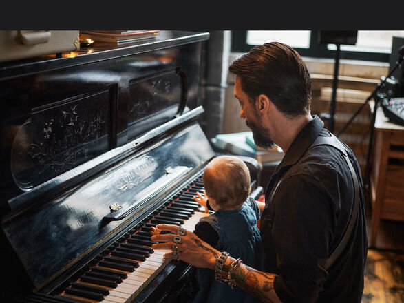 Musician Alec Voelkel sits at a piano with a toddler on his lap, playing in a warmly furnished room
