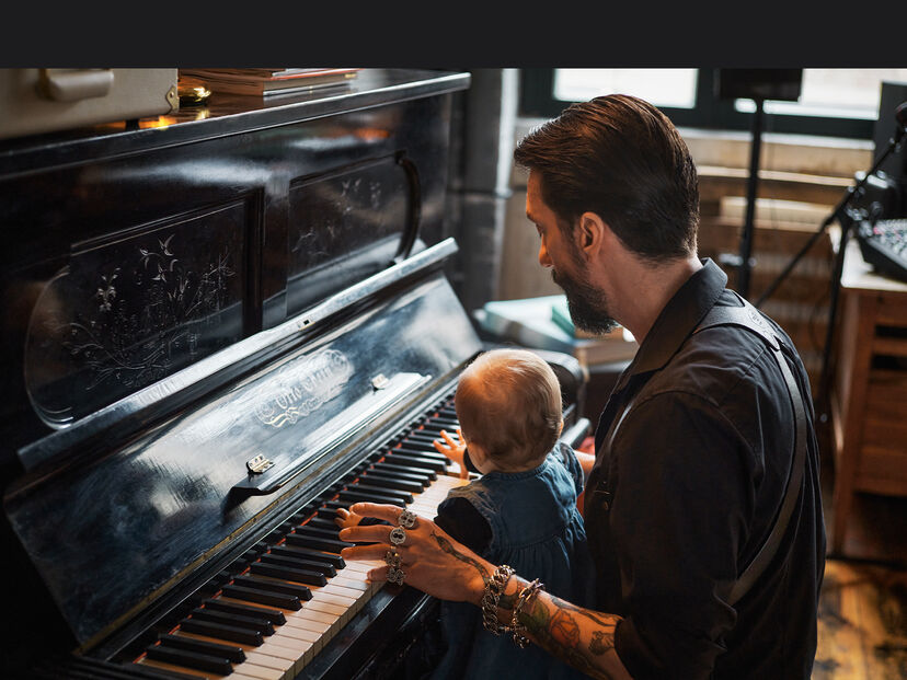 Der Musiker Alec Voelkel sitzt mit einem Kleinkind auf dem Scho&szlig; am Klavier und spielt, in einem gem&uuml;tlich eingerichteten Raum