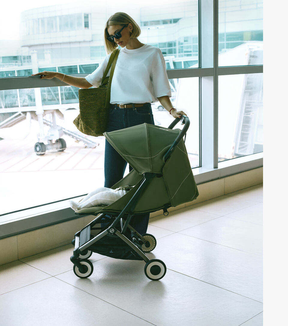 A woman with sunglasses and a bag stands next to the Orfeo compact travel stroller at an airport