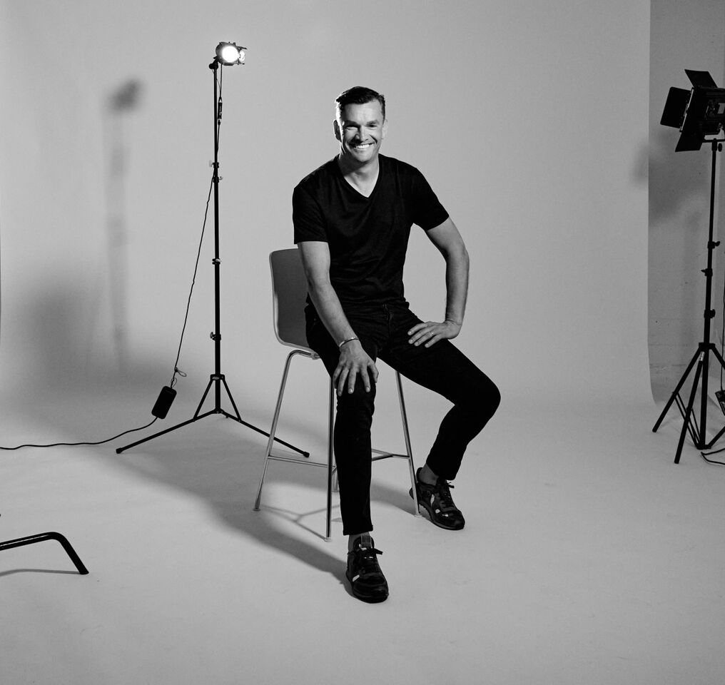 A man smiling while sitting on a chair in a photo studio, surrounded by lighting equipment.
