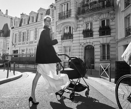 A woman in an elegant dress and coat walks with a stroller on a Parisian street.