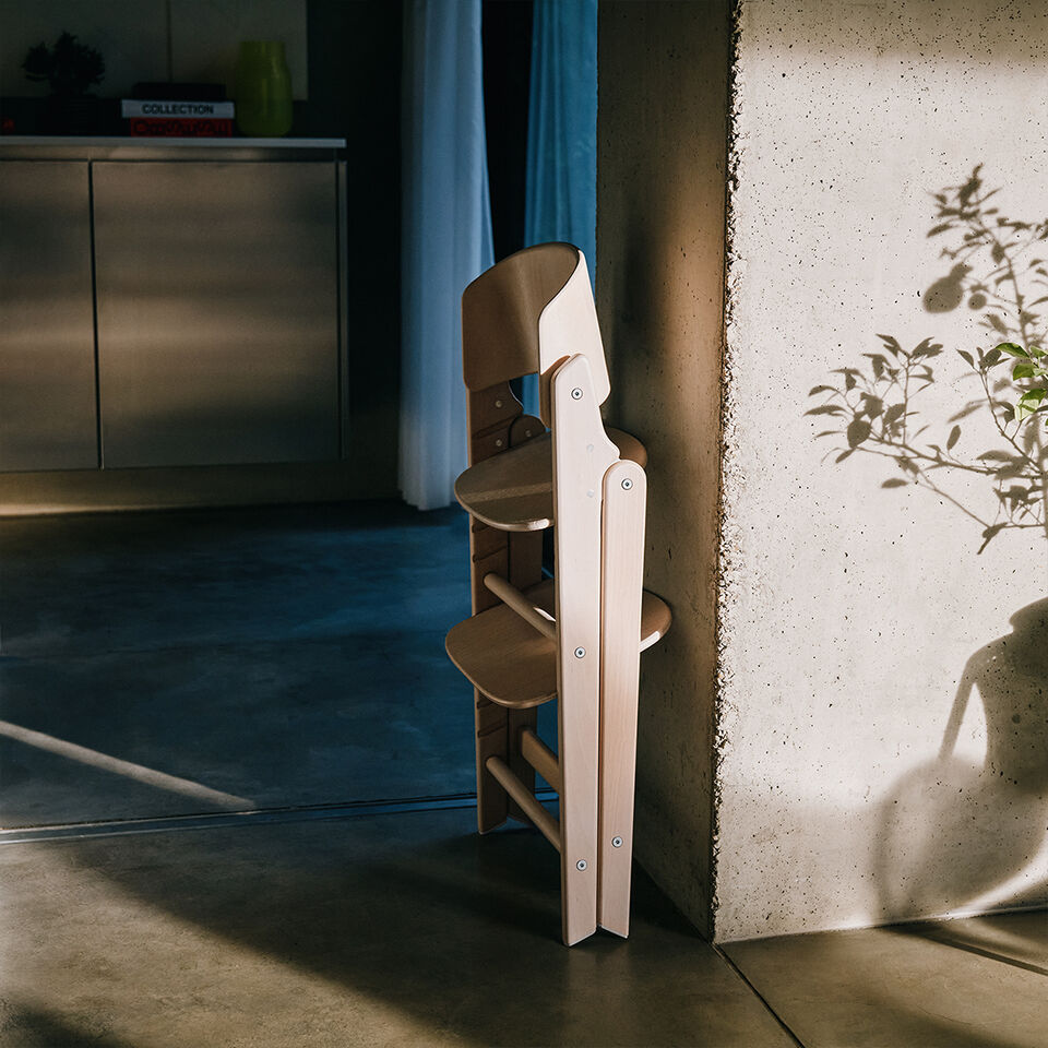 Click and Fold wooden high chair, folded and leaning against a wall.