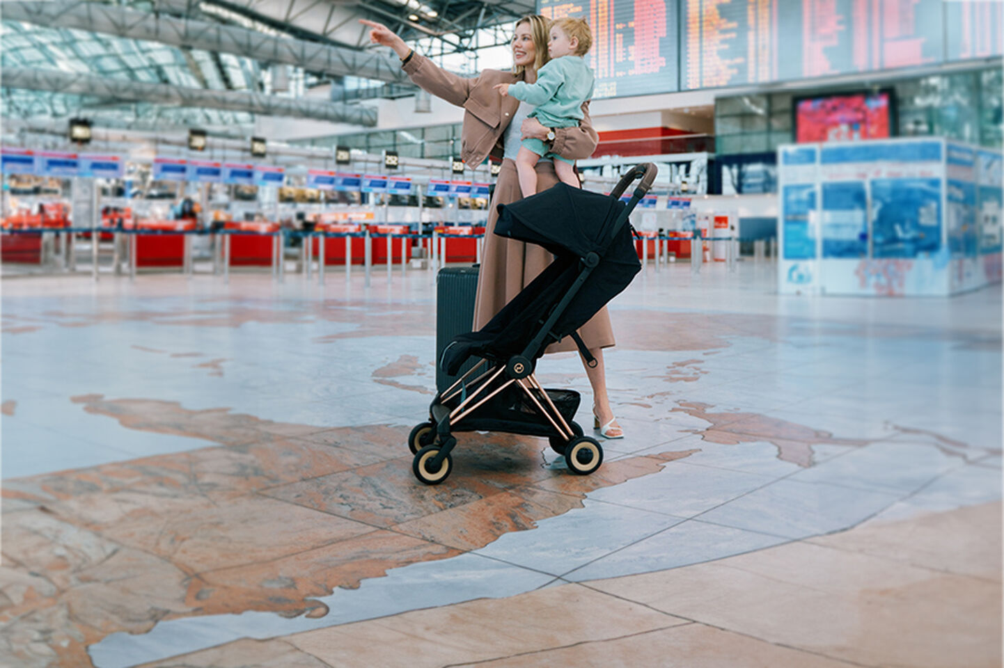 A woman holding a child points at something in an airport, with a black stroller beside them.