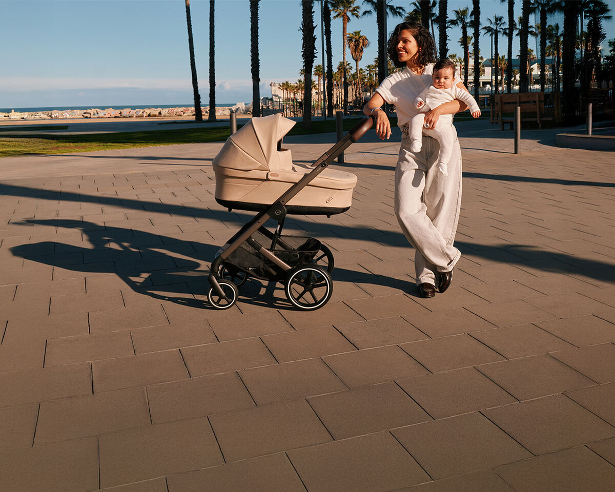 A woman standing with her arm leaning against the handlebar of her Balios S Lux stroller. With her other hand, she's holding her baby.