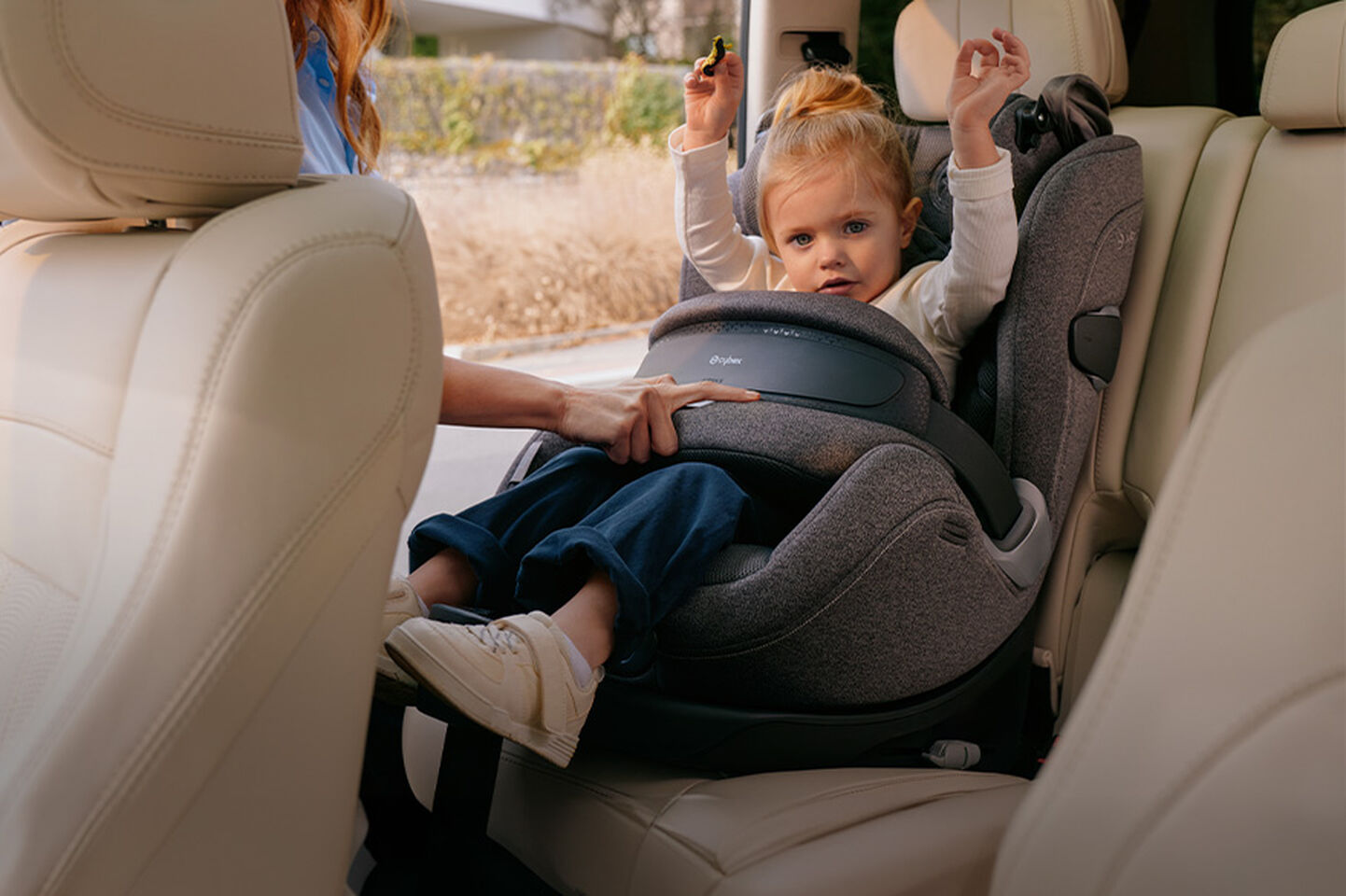 A small child sits in a car seat in the backseat of a car while an adult adjusts the seat.