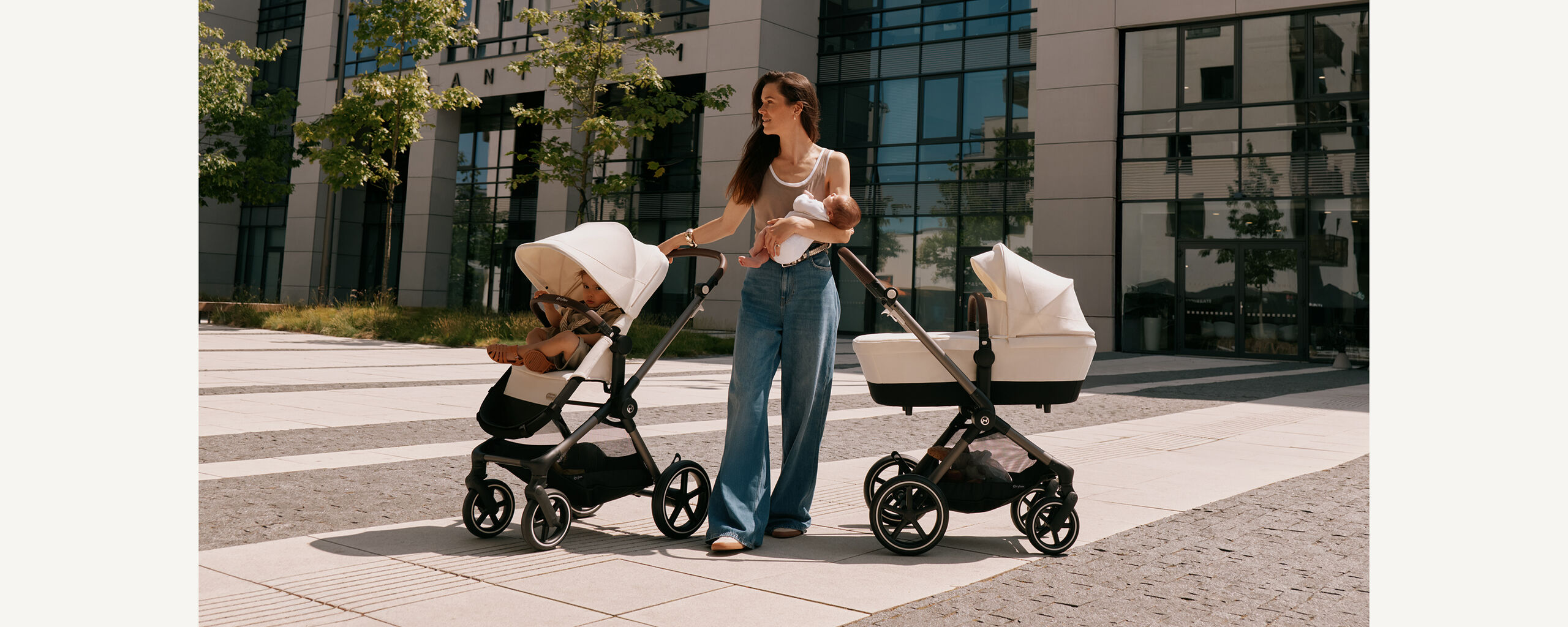 CYBEX-barnvagnsduo &ndash; en mamma h&aring;ller sin bebis i famnen. Bredvid henne st&aring;r tv&aring; barnvagnar utomhus, en med ett s&auml;te f&ouml;r sm&aring;barn och en med en liggdel i en solig stadsmilj&ouml;