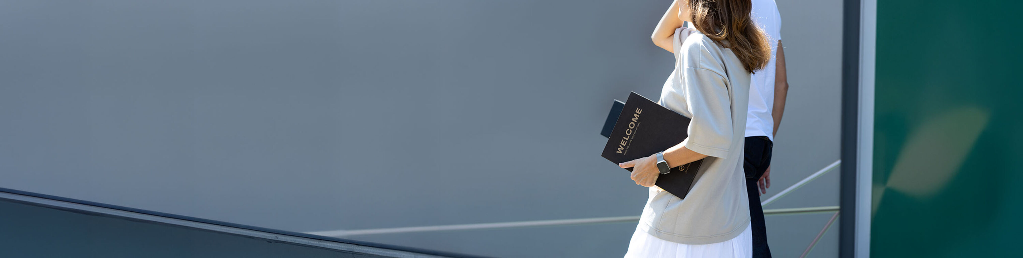 Cropped rear view of a woman and a man walking outdoors; the woman carries a folder labeled &ldquo;Welcome.&rdquo;
