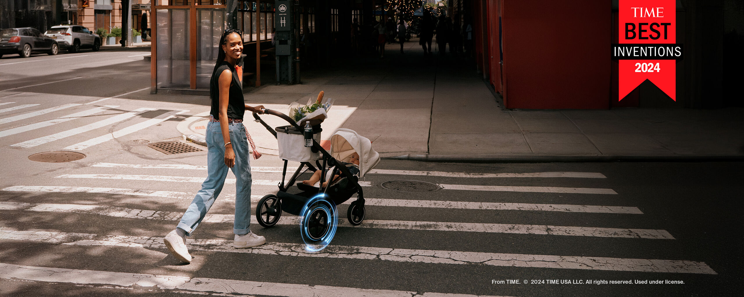 A woman with long hair walks with a stroller across a crosswalk in a city, surrounded by modern buildings.