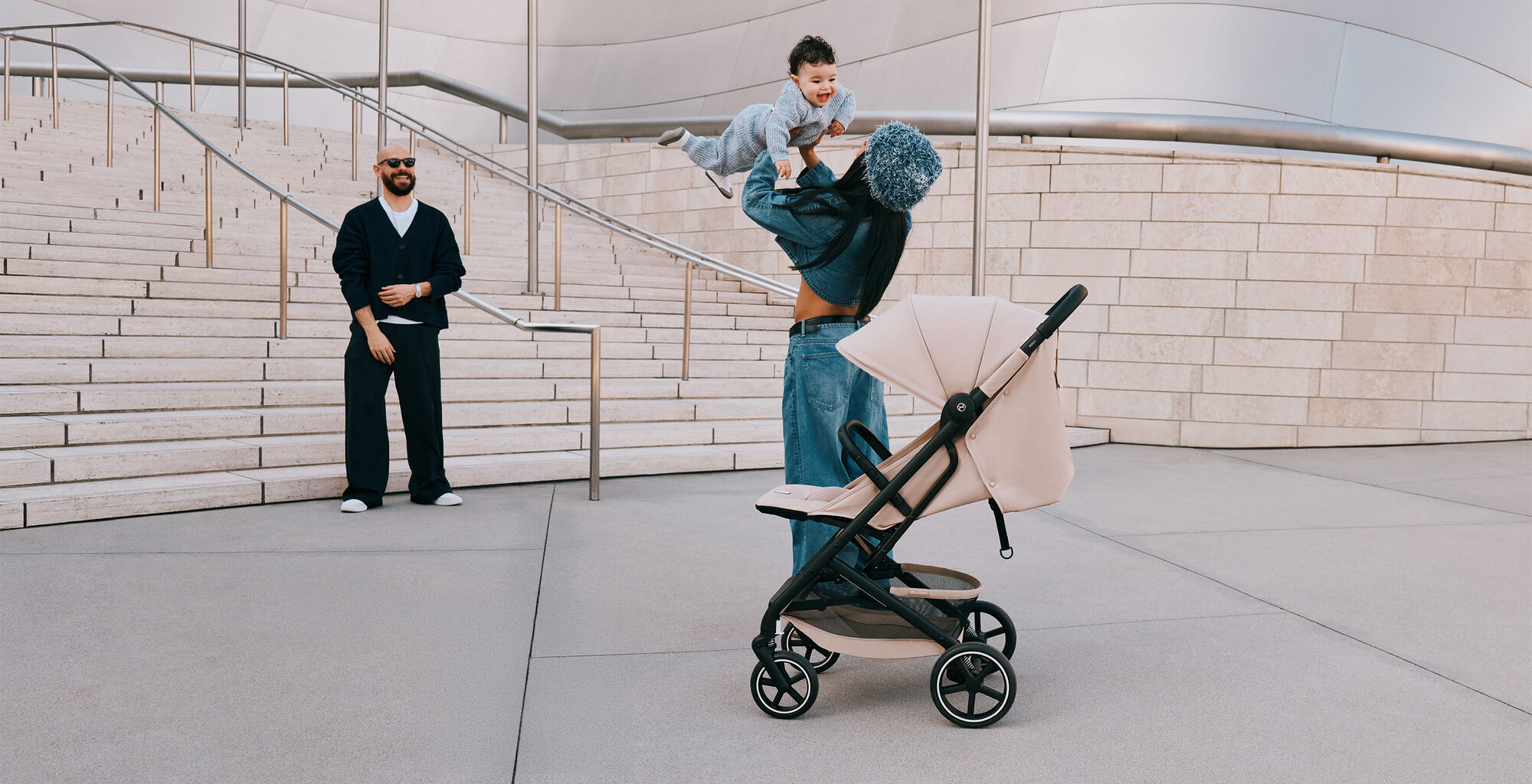 Une femme joue avec son fils en le lan&ccedil;ant doucement en l&rsquo;air, tandis que le p&egrave;re les observe en riant. Une poussette compacte Beezy se tient devant eux.